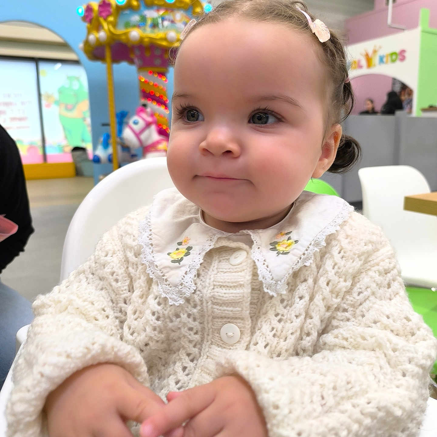 Nélia participe au concours pour gagner de l'argent avec cette photo : toddler, child, indoor, high_chair, knitted_sweater, curly_hair, hair_clip, hands, face, expression, playground, carousel, colorful, background, seated, cute, portrait, person, clothing, baby