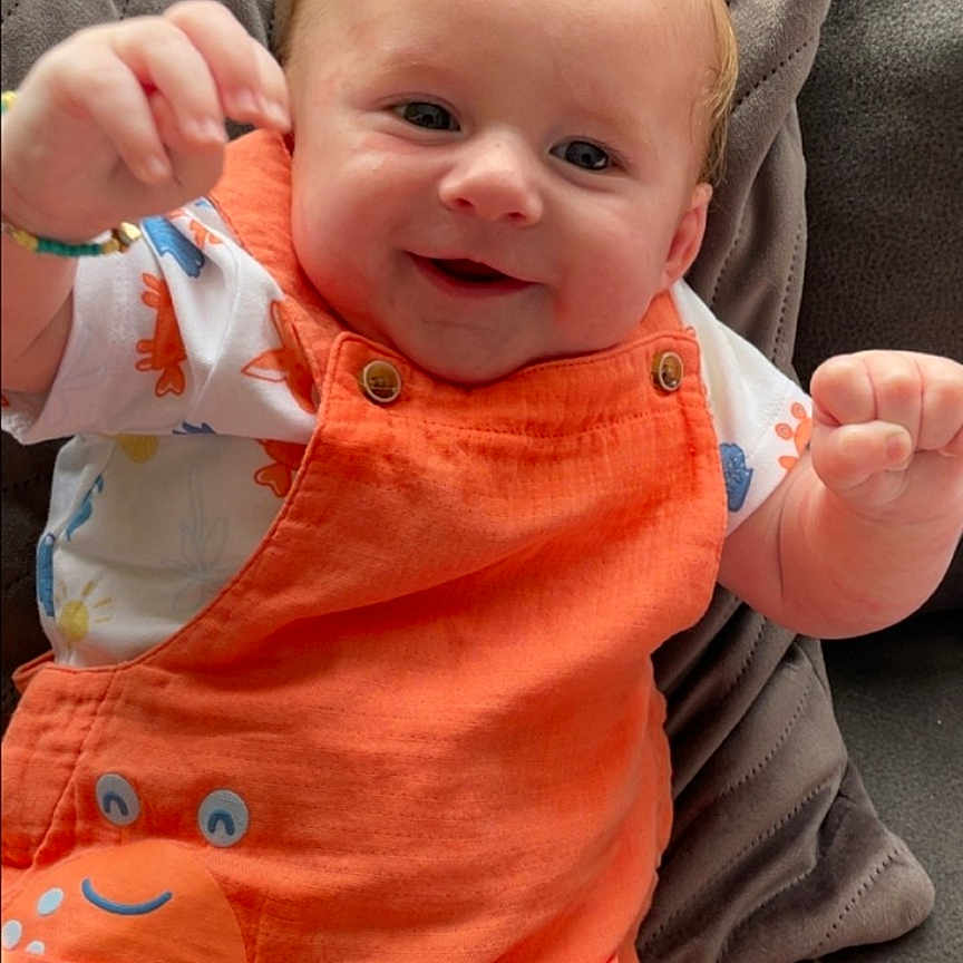 Lyam participe au concours pour gagner de l'argent avec cette photo : baby, casual, child, clothing, comfort, couch, cute, face, furniture, happy, head, indoor, infant, orange_overalls, person, pillow, plush, sitting, smiling, young_child