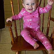 Kodi Sue is registered to the contest to win money with this photo: toddler, child, smiling, pajamas, pink_clothing, wooden_chair, rocking_chair, indoor, tile_floor, happy, waving, person, cute, baby, sitting, face, small_child, home, furniture, joyful