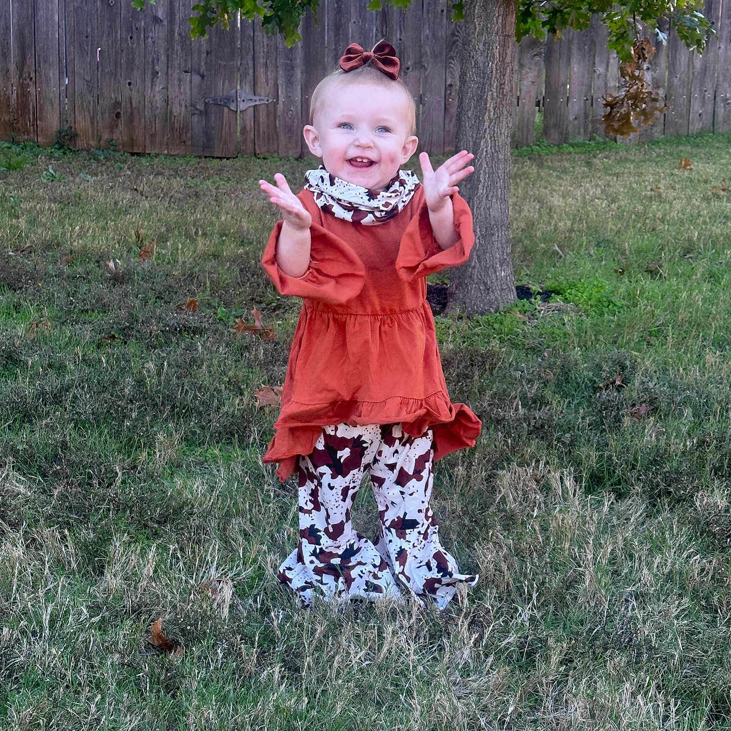 Kodi Sue is registered to the contest to win money with this photo: toddler, child, smiling, outdoor, grass, tree, wooden_fence, dress, patterned_pants, bow, happy, clapping, person, nature, daylight, greenery, playful, young, fashion, portrait