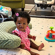 Journey is registered to the contest to win money with this photo: baby, child, infant, pink_dress, polka_dots, bow, afro_hair, play_mat, toy, pillow, cushion, indoor, floor, carpet, smile, sitting, cute, home, furniture, happy