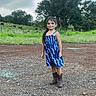 child, girl, blue_dress, boots, outdoor, standing, gravel, nature, trees, cloudy_sky, smiling, long_hair, field, grass, young, person, casual_clothing, daytime, rural, portrait