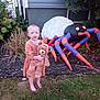 child, toddler, barefoot, teddy_bear, orange_dress, grass, garden, inflatable_spider, decorations, house, plants, shrubbery, fall_leaves, outdoor, cute, toy, lighting, window, hedge, yard