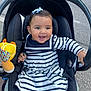 baby, black_car_seat, blue_bow, car_seat, child, cute, face, hand, happy, infant, outdoor, pavement, person, plush_toy, purple_tights, road, seatbelt, smiling, striped_dress, toy
