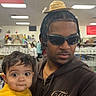 adult, baby, background, braids, casual_clothing, child, cute, expression, glasses, hat, holding, indoor, man, people, person, portrait, shopping, store, sunglasses, yellow_sweater