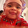 Kayden is registered to the contest to win money with this photo: baby, child, face, tongue_out, bonnet, red_clothing, indoor, closeup, person, portrait, funny_expression, headwear, cute, smiling, human, young_child, warm_colors, wall_art, blurred_foreground, background_person