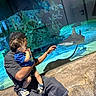 adult, aquarium, blue, casual_clothing, child, curious, exhibit, fish, glass, hand, holding, indoor, marine_life, people, reflection, rocks, shark, swimming, tank, water