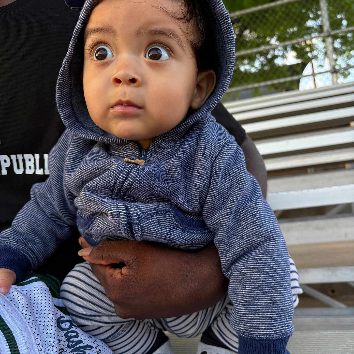 Kayden joined the competition — help win amazing prizes! baby, child, hoodie, blue_clothing, mickey_mouse, socks, striped_pants, pointing, wide_eyes, outdoor, bench, adult_hand, black_skin, curious_expression, metal_bleachers, green_foliage, casual_clothing, portrait, daylight, cute