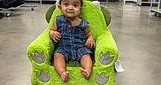 Kayden joined the competition — help win amazing prizes! baby, bag, child, childrens_clothing, concrete_floor, curious, door, floor, green_chair, indoor, person, pillows, plush_chair, retail, seated, shelves, smiling, sports_equipment, store, toy_chair