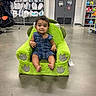 baby, bag, child, childrens_clothing, concrete_floor, curious, door, floor, green_chair, indoor, person, pillows, plush_chair, retail, seated, shelves, smiling, sports_equipment, store, toy_chair