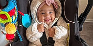 Yvanah participe au concours pour gagner de l'argent avec cette photo : baby, child, car_seat, smiling, toy, headband, pink_bow, fluffy_jacket, indoor, floor, black_seat, white_socks, pacifier, colorful_toys, happy, infant, seated, clothing, safety_harness, cute