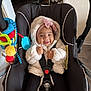 baby, child, car_seat, smiling, toy, headband, pink_bow, fluffy_jacket, indoor, floor, black_seat, white_socks, pacifier, colorful_toys, happy, infant, seated, clothing, safety_harness, cute