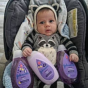 Luciano is registered to the contest to win money with this photo: baby, infant, knitted_clothing, raccoon_outfit, baby_carrier, blanket, johnsons, baby_bath_products, purple_bottles, cute, wide_eyes, indoors, flooring, child, person, portrait, soft_toys, cozy, warm_clothing, newborn
