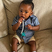 Regis is registered to the contest to win money with this photo: toddler, child, baby, pacifier, blue_shirt, shorts, white_shoes, couch, indoor, sitting, curly_hair, brown_skin, clothing, footwear, comfort, cute, portrait, young_child, toy, relaxed