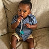 toddler, child, baby, pacifier, blue_shirt, shorts, white_shoes, couch, indoor, sitting, curly_hair, brown_skin, clothing, footwear, comfort, cute, portrait, young_child, toy, relaxed
