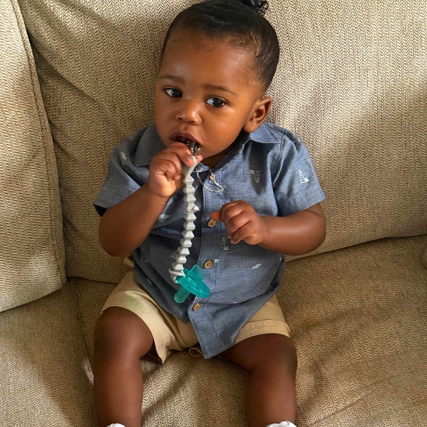 Regis is registered to the contest to win money with this photo: toddler, child, baby, pacifier, blue_shirt, shorts, white_shoes, couch, indoor, sitting, curly_hair, brown_skin, clothing, footwear, comfort, cute, portrait, young_child, toy, relaxed