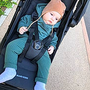 Alex a rejoint le concours — aidez-le/la à gagner de superbes lots ! baby, stroller, child, outdoor, pavement, hat, clothing, footwear, green_outfit, black_stroller, baby_wear, infant, person, seat_belt, casual, daylight, sidewalk, relaxed, baby_sock, baby_hat