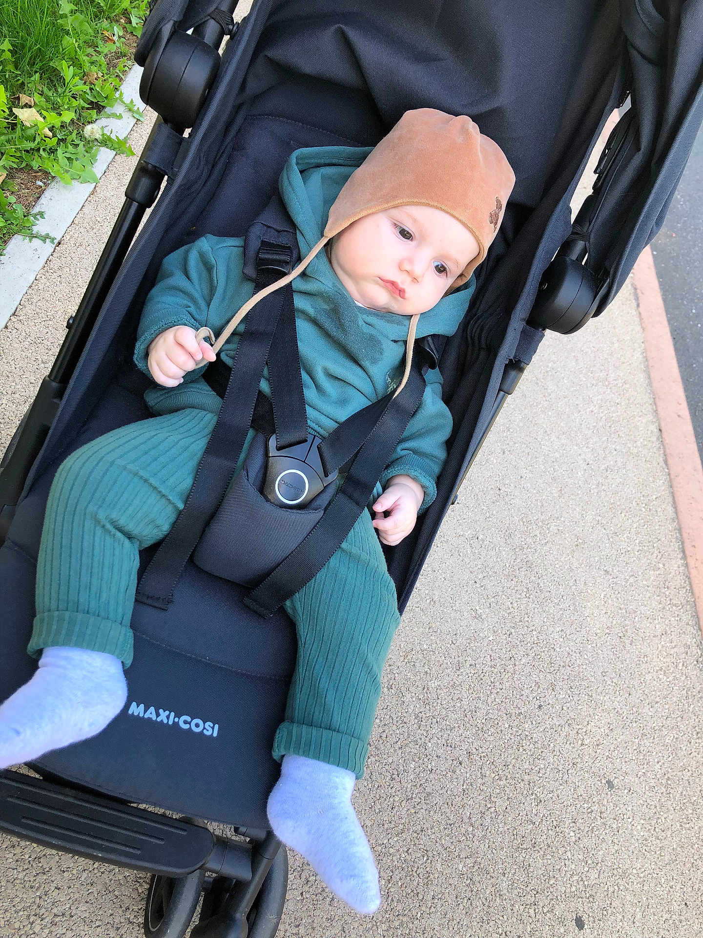 Alex a rejoint le concours — aidez-le/la à gagner de superbes lots ! baby, stroller, child, outdoor, pavement, hat, clothing, footwear, green_outfit, black_stroller, baby_wear, infant, person, seat_belt, casual, daylight, sidewalk, relaxed, baby_sock, baby_hat