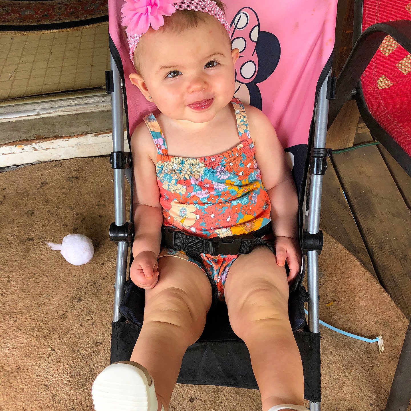 Zarra is registered to the contest to win money with this photo: baby, carpet, child, cute, floral_outfit, flower_headband, footwear, happy, headwear, outdoor, person, pink, playful, sandals, seat, sitting, smiling, stroller, toddler, wooden_floor