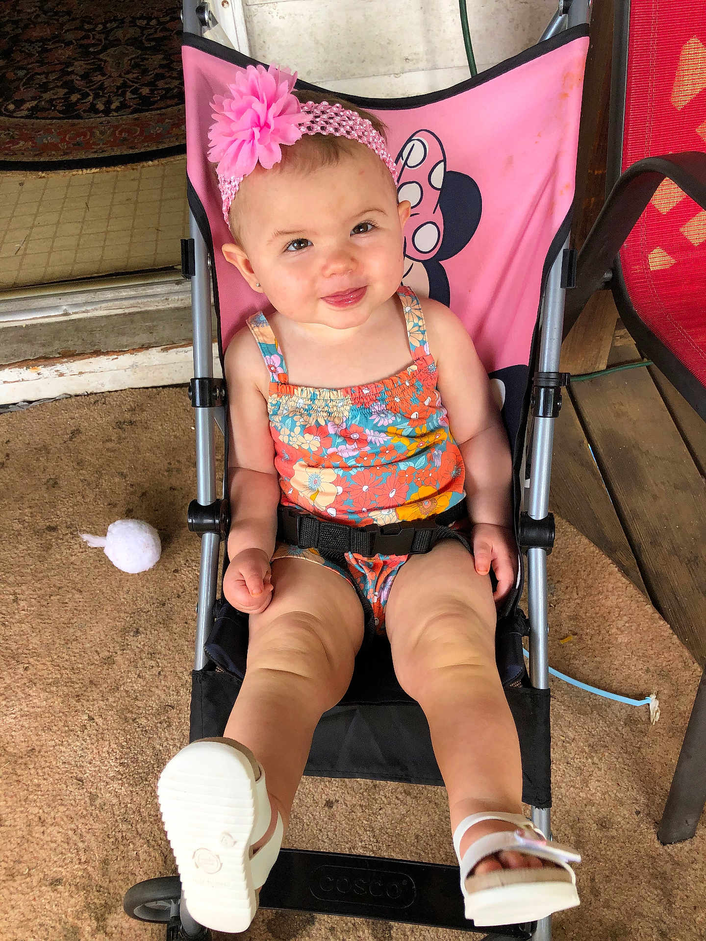 Zarra is registered to the contest to win money with this photo: toddler, child, stroller, pink, flower_headband, smiling, sitting, outdoor, sandals, floral_outfit, baby, cute, playful, person, footwear, headwear, happy, carpet, wooden_floor, seat