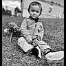 toddler, child, outdoor, grass, flowers, black_and_white, building, casual_clothing, hoodie, pants, shoes, serious_expression, portrait, nature, daylight, sitting, young_child, face, person, photography