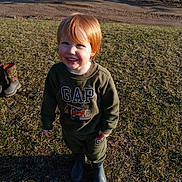 Landon is registered to the contest to win money with this photo: child, smiling, outdoor, grass, green_clothing, red_hair, boots, sunlight, happy, toddler, sweatshirt, pants, person, nature, daylight, casual_clothing, playful, cute, young, portrait