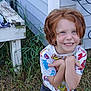 child, red_hair, smiling, outside, grass, shirt, cartoon_print, house_siding, wooden_table, playful, happy, person, shorts, nature, young, cute, portrait, fun, daylight, expression
