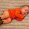 baby, blanket, brown_shoes, child, closed_eyes, cozy, curled_up, face, indoor, infant, lying_down, orange_clothing, peaceful, photography, portrait, sleeping, smiling, striped_pants, textured_fabric, warm_colors