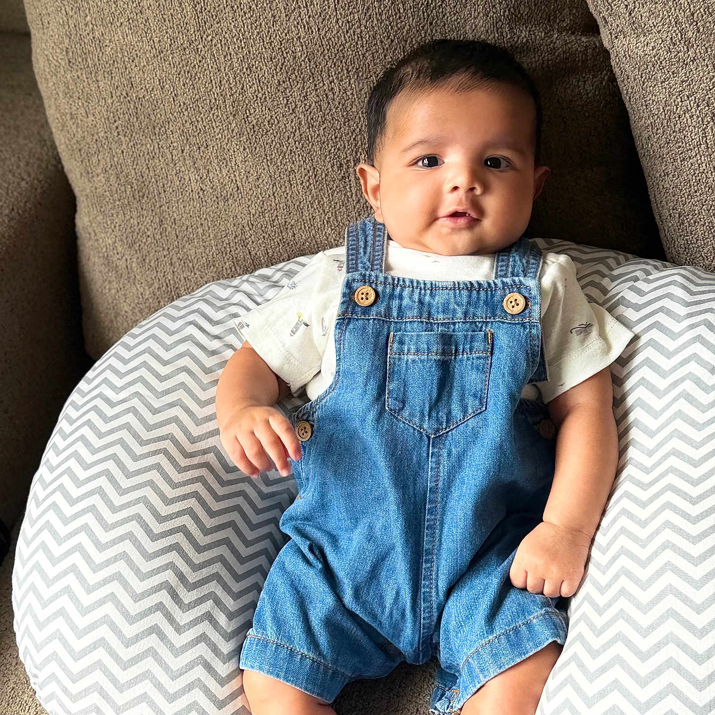 Amir Gaffar is registered to the contest to win money with this photo: baby, denim_overalls, white_tshirt, chevron_pillow, brown_couch, infant, sitting, indoor, cute, child, smiling, face, person, footwear, white_socks, plush, comfortable, portrait, young_child, casual_clothing