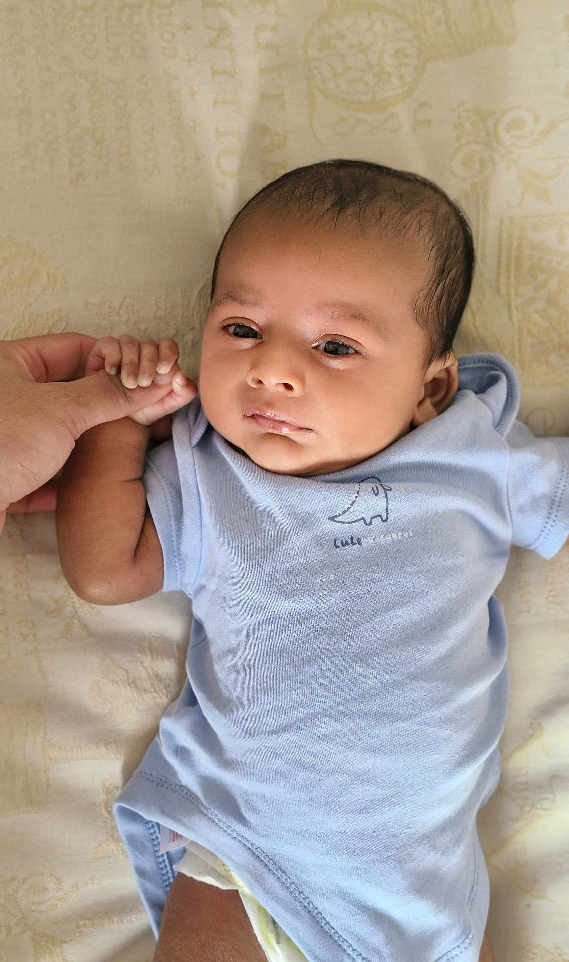 Ivaan is registered to the contest to win money with this photo: baby, infant, child, hand, finger, onesie, blue_clothing, dinosaur_print, blanket, bed, lying_down, skin, face, eyes, person, newborn, cute, tender, soft, indoors