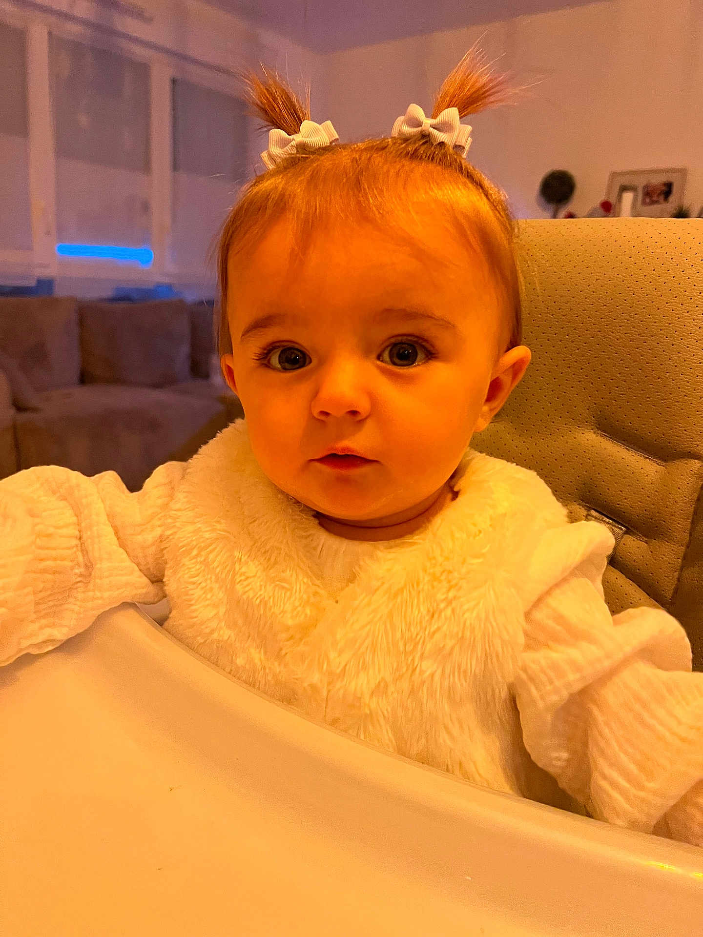 Romy a rejoint le concours — aidez-le/la à gagner de superbes lots ! baby, child, pigtails, white_bow, high_chair, white_clothing, curious, indoor, living_room, sofa, window, soft_lighting, face, portrait, cute, young_child, infant, person, seated, cozy