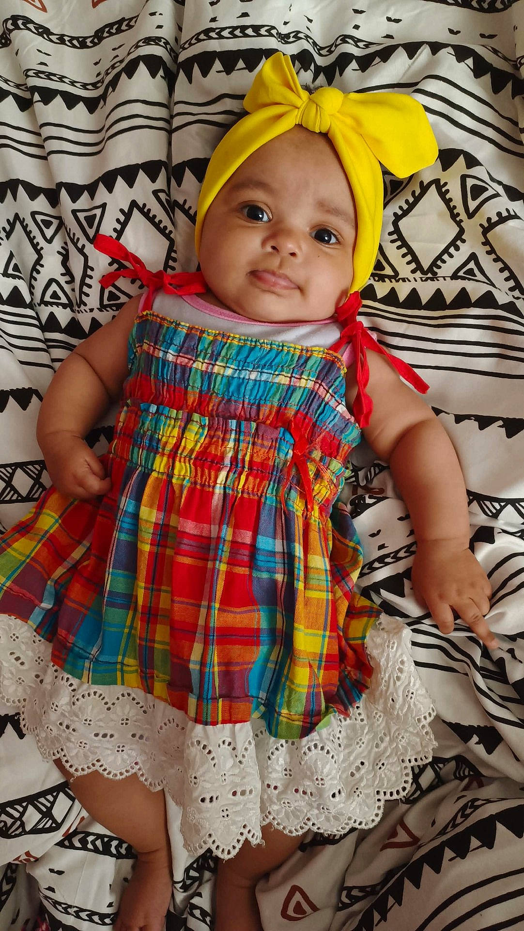 Jah' a rejoint le concours — aidez-le/la à gagner de superbes lots ! arms, baby, blanket, child, colorful_dress, curious_expression, cute, face, headwear, indoors, infant, lace_trim, legs, lying_down, patterned_blanket, plaid_pattern, portrait, skin, smile, yellow_headband