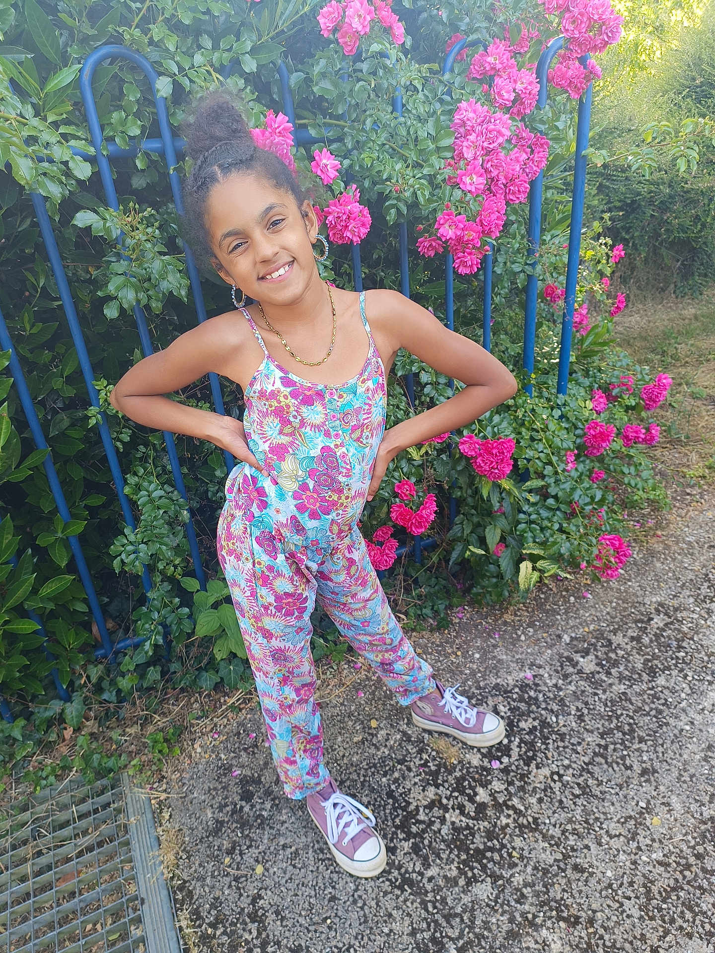 Naï participe au concours pour gagner de l'argent avec cette photo : child, girl, smiling, standing, outdoor, flowers, pink_flowers, greenery, blue_fence, jumpsuit, colorful_clothing, sneakers, hair_bun, earrings, necklace, nature, sunlight, pathway, plants, casual