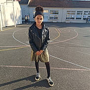 Naï participe au concours pour gagner de l'argent avec cette photo : child, girl, basketball_court, school, outdoor, sunny, black_jacket, gold_skirt, sneakers, headband, smile, standing, asphalt, trees, building, windows, shadow, playground, casual_clothing, young