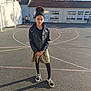 asphalt, basketball_court, black_jacket, building, casual_clothing, child, girl, gold_skirt, headband, outdoor, playground, school, shadow, smile, sneakers, standing, sunny, trees, windows, young