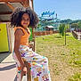 child, girl, curly_hair, orange_top, floral_skirt, sandals, wooden_railing, amusement_park, bobsleigh, outdoor, sunny, grass, tree, people, blue_sky, smile, casual_clothing, daytime, fence, recreation