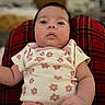 baby, infant, child, floral_clothing, pink_clothing, plaid_blanket, red_blanket, face, eyes, arm, indoors, blurred_background, cute, expression, newborn, skin, person, lying_down, portrait, soft_lighting