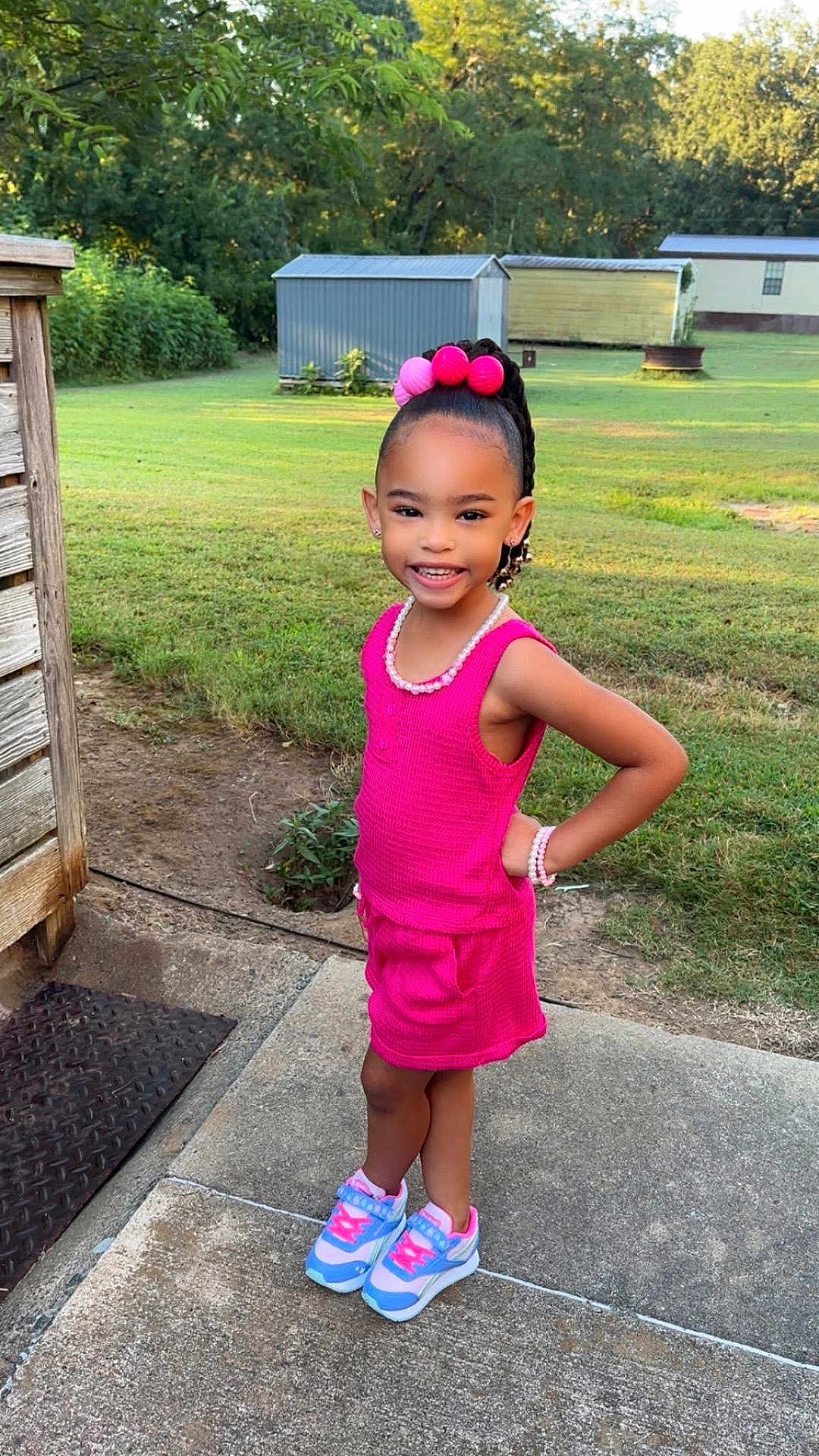 Ava joined the competition — help win amazing prizes! child, girl, outdoor, grass, concrete, sidewalk, pink_clothing, sneakers, bracelet, necklace, hair_accessory, smiling, standing, earrings, pose, sunny, daylight, yard, shed, nature
