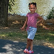 Ava is registered to the contest to win money with this photo: child, girl, smiling, pose, outdoor, lake, tree, grass, sidewalk, sandals, shorts, striped_shirt, sunlight, shadow, nature, happy, summer, playful, casual_clothing, daytime