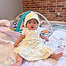 baby, infant, child, dress, bonnet, quilt, toy, playmat, floor, brick_wall, indoor, cute, sitting, chubby_cheeks, colorful, pattern, person, headwear, foot, smiling