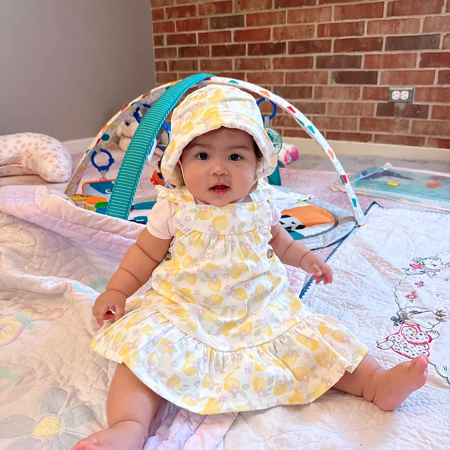 Lily joined the competition — help win amazing prizes! baby, infant, child, dress, bonnet, quilt, toy, playmat, floor, brick_wall, indoor, cute, sitting, chubby_cheeks, colorful, pattern, person, headwear, foot, smiling