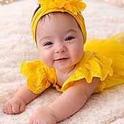 Ainhoa is registered to the contest to win money with this photo: baby, infant, smiling, yellow_dress, headband, flower, blanket, soft_texture, cute, portrait, happy, lying_down, indoors, child, toddler, bright_colors, warm_lighting, bokeh, close_up, adorable