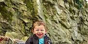 Aloïs a rejoint le concours — aidez-le/la à gagner de superbes lots ! toddler, child, smiling, barefoot, jacket, red_pants, wooden_bench, rocky_background, outdoor, nature, happy, portrait, sitting, casual_clothing, person, baby, cute, fun, daylight, young_child