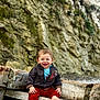 toddler, child, smiling, barefoot, jacket, red_pants, wooden_bench, rocky_background, outdoor, nature, happy, portrait, sitting, casual_clothing, person, baby, cute, fun, daylight, young_child