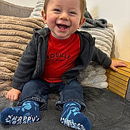 Aloïs a rejoint le concours — aidez-le/la à gagner de superbes lots ! toddler, child, smiling, happy, red_shirt, hoodie, socks, jeans, couch, pillows, indoor, cute, baby, seated, cozy, wooden_armrest, face, person, casual_clothing, home