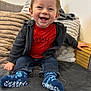 toddler, child, smiling, happy, red_shirt, hoodie, socks, jeans, couch, pillows, indoor, cute, baby, seated, cozy, wooden_armrest, face, person, casual_clothing, home