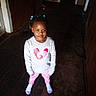 braided_hair, child, dark_interior, full_body, girl, hair_beads, hallway, heart_design, home, indoors, leggings, pink_pants, portrait, smiling, socks, standing, sweatshirt, toddler, wooden_floor, wooden_walls