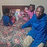 adult, birthday, blue_hoodie, candy, carpet, child, children, family, floral_pattern, furniture, gathering, grainy_photo, group_photo, indoor, living_room, lollipop, pink_shirt, portrait, sitting, snacks