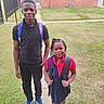 children, siblings, school, backpacks, outdoors, portrait, standing, smiling, braids, school_uniform, polo_shirt, red_shirt, black_clothing, sneakers, pink_shoes, grass, brick_building, sidewalk, fence, daylight
