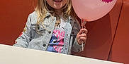 Giulia a rejoint le concours — aidez-le/la à gagner de superbes lots ! balloon, blonde_hair, booth, cartoon_character, casual_clothing, celebration, child, denim_jacket, fun, girl, happy, holding, indoor, person, pink, portrait, restaurant, seated, smile, table